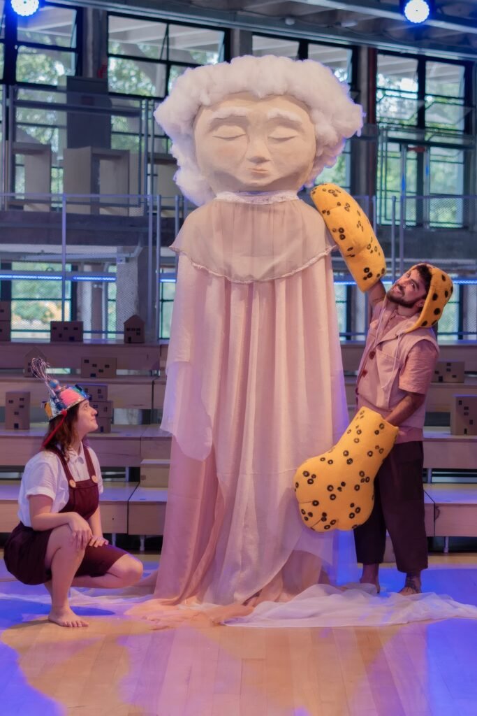 Boneco gigante da Velha Ventania, figura de véu branco ladeada por dois atores em cena de Do Outro Lado do Rio