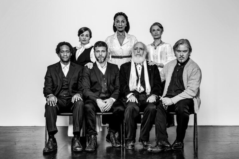 Retrato em preto e branco do elenco de O Dilema de Sidgwick, sete atores sentados e em pé contra fundo claro